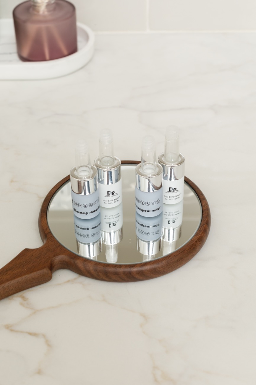 Skincare vials arranged on a wooden hand mirror on a marble counter